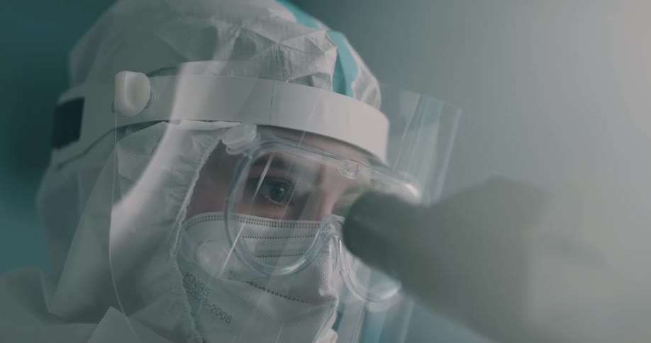 A lab worker wearing protective clothing and a plastic shield looks into a microscope