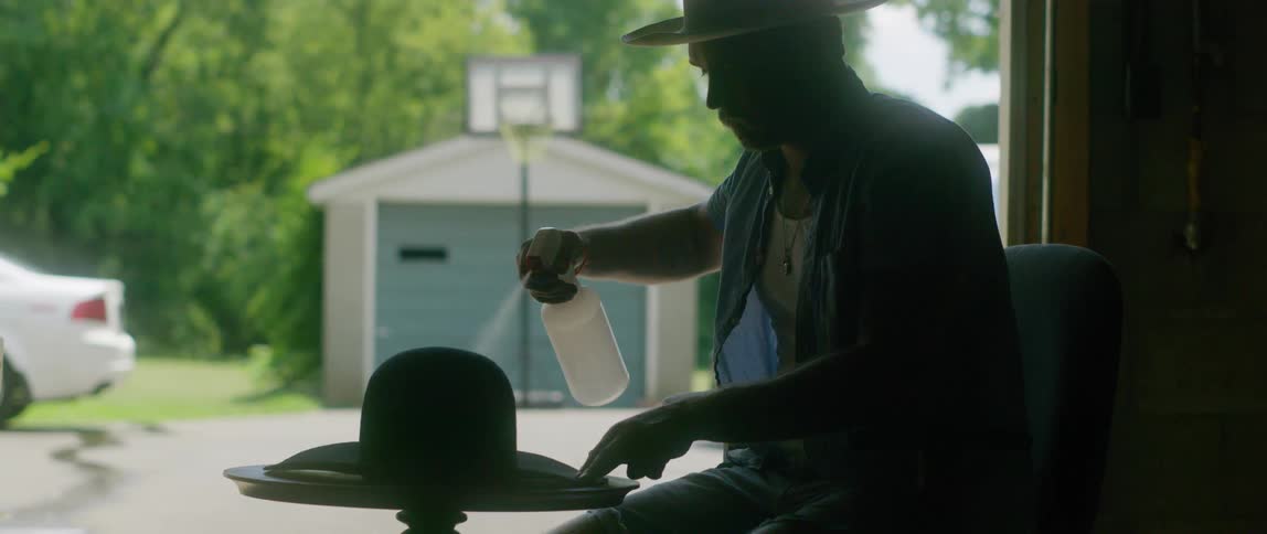 A man with a hat sitting on a chair spraying a hat with a spray bottle