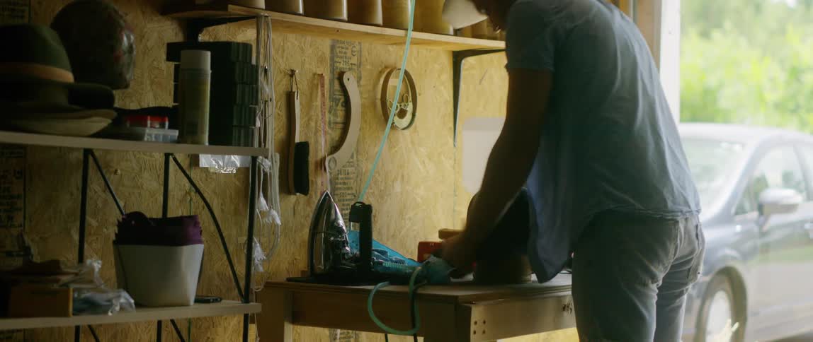 Man works on making hat in open door workshop while dark colored car sits parked nearby