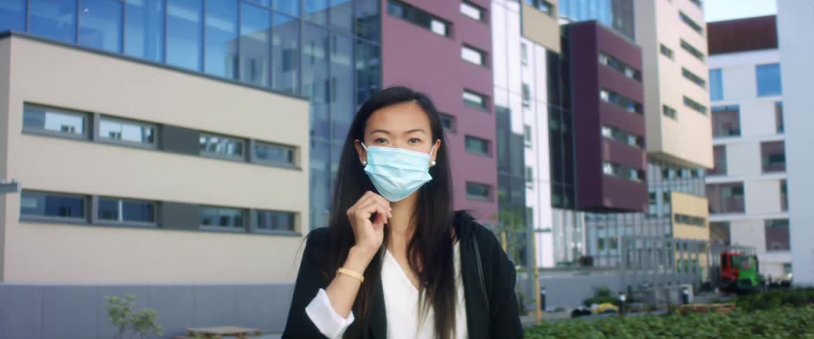 Woman looking at camera puts a facemask on her face while standing next to buildings in the city