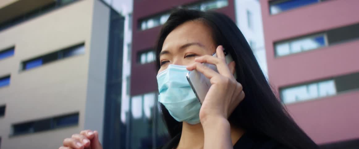 A woman standing outdoors in a medical mask wags her finger as she talks on her smartphone