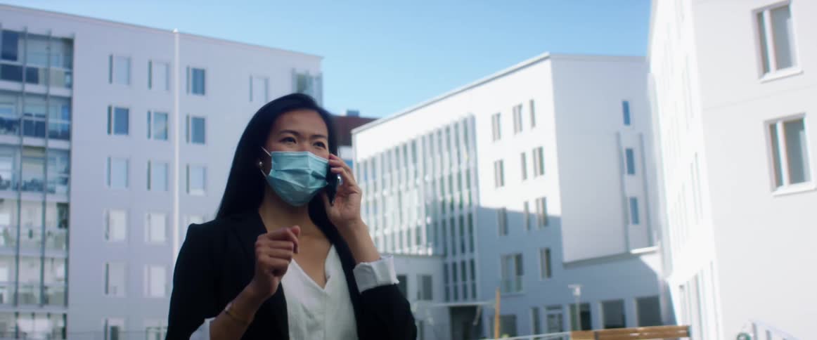 Woman stands outdoors nearlow rise buildings wearing face mask gesturing as she talks on mobile phone