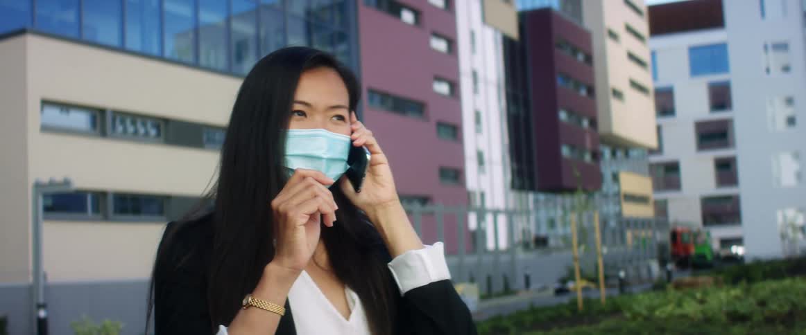 A woman stands outside a large city building talking on her cell phone and wearing a face mask