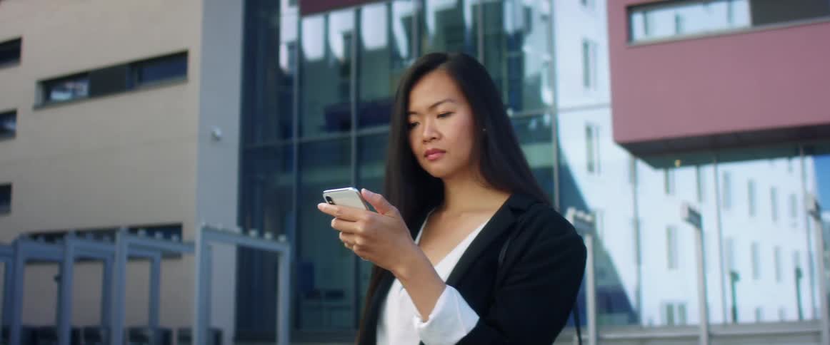 Asian woman with long hair a black blazer and a white shirt uses iphone xs in front of a building in the street