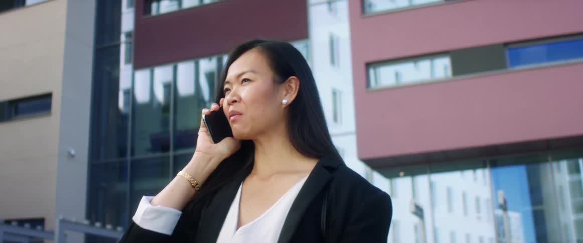 Asian woman with long straight hair a black blazer and a white shirt talks on the smartphone in the city street with high rise buildings in the background