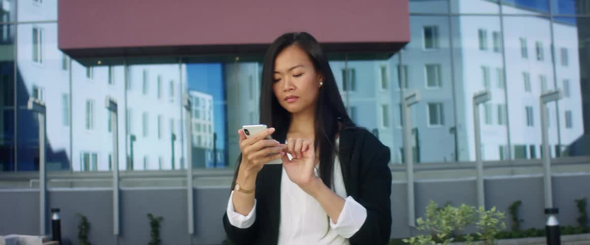 Asian woman with long straight hair uses an iphone smartphone in frontof a high rise building