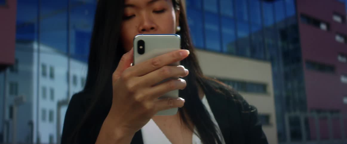 Asian woman with long hair and a black blazer uses an iphone in the city street