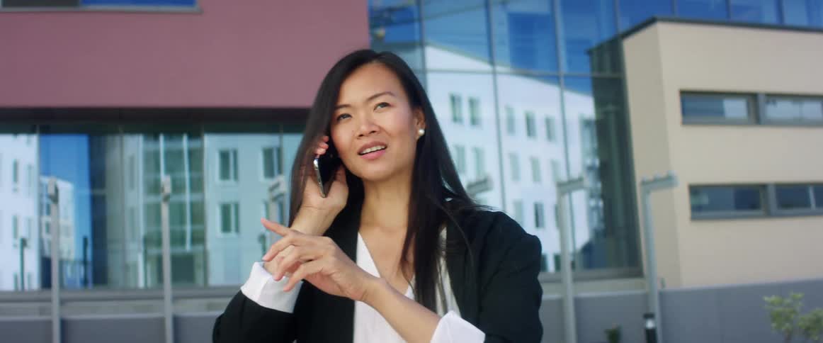 Asian woman with long hair and black blazer talks on smartphone in front of high rise buildings