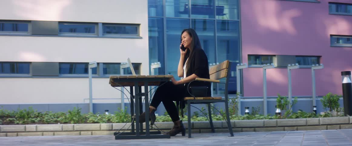 An asian woman talks on a smartphone while sitting at a small table outdoors