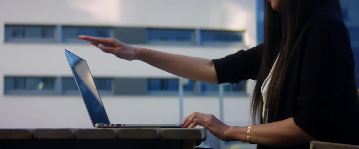 Asian woman with a white shirt and a black jacket sits at a wood table and opens a laptop to use outdoors