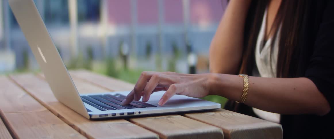 A woman types on a laptop with one hand while sitting at a wooden table outdoors during the day