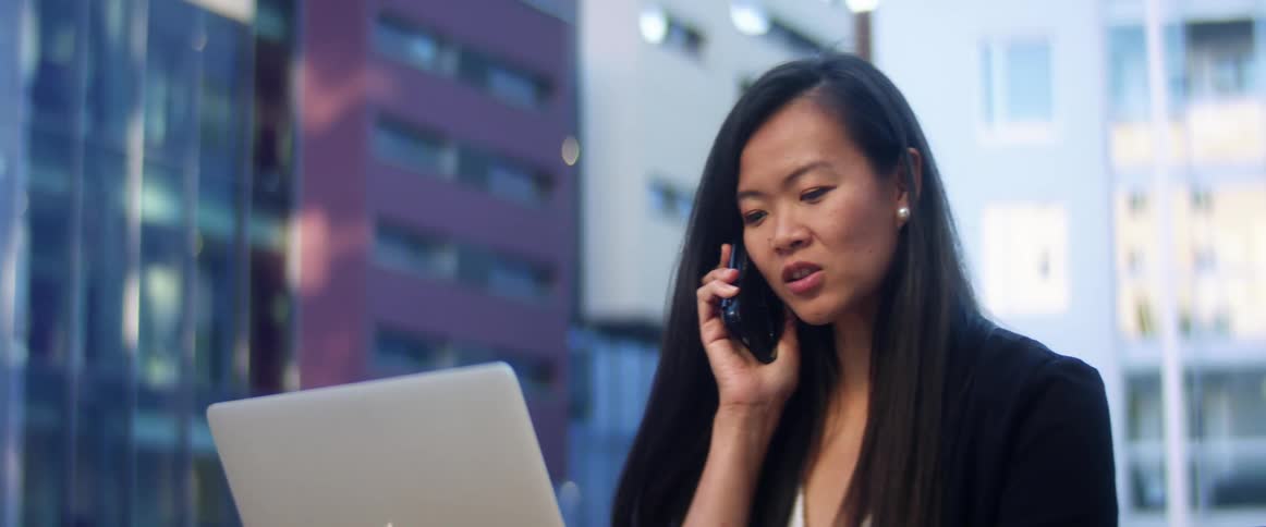 An asian woman talks on a smartphone while she sits in front of a laptop at a table outdoors in a metropolis during the day