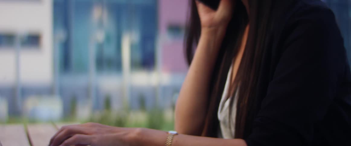 Asian woman with a long straight hair and a blazer sits at a wood table and talks on the smartphone while using a grey laptop