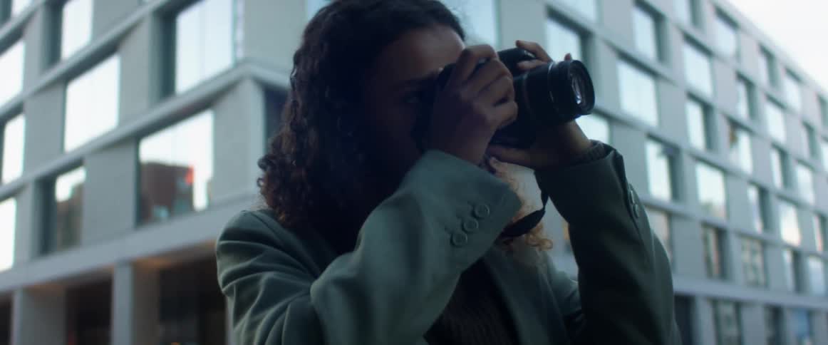 A young female photographer standing outdoors in a city raises her camera to take a photograph