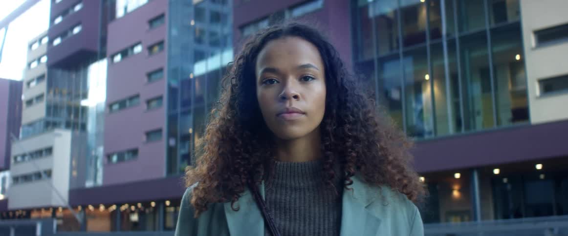Black woman with curly hair and a light blue coat faces the camera with eye contact in the city street with buildings in the background