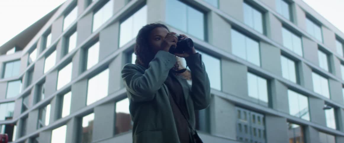Black woman with curly hair and a blue top coat takes photographs and checks them in the street