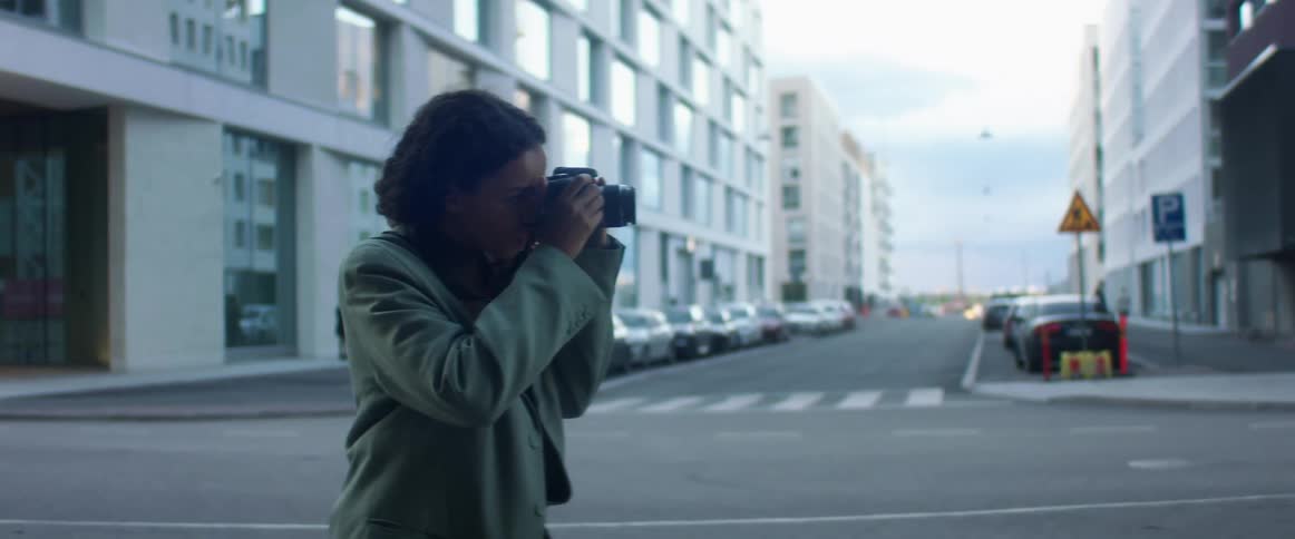 A woman stands near a quiet city intersection and takes a photo with a professional camera
