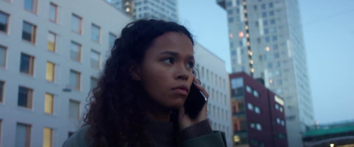 Black woman with curly hair talking on the smartphone looks worried in city street with high rise buildings in the background