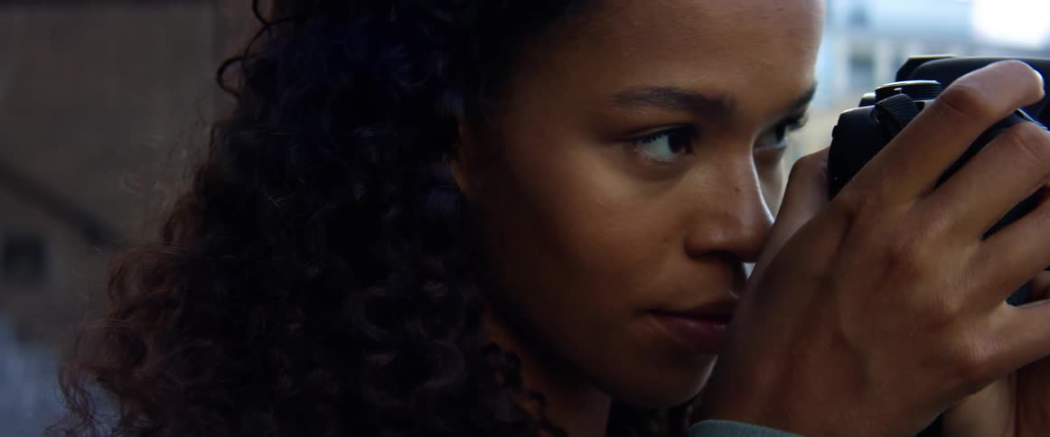 Black woman with curly hair and a blue top coat takes photographs and checks them in a street