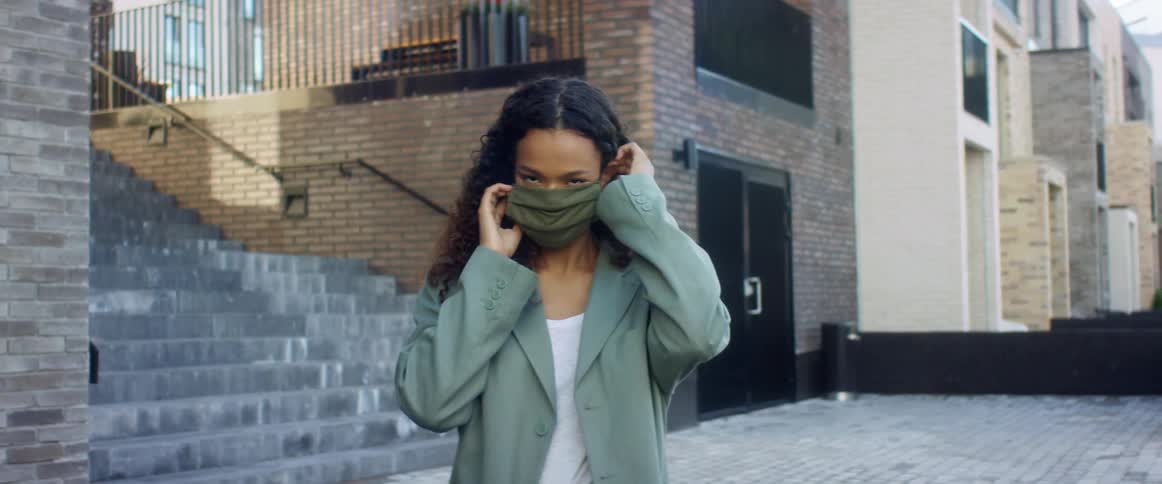 Black woman with curly hair and a blue overcoat wears a grey mask in the street