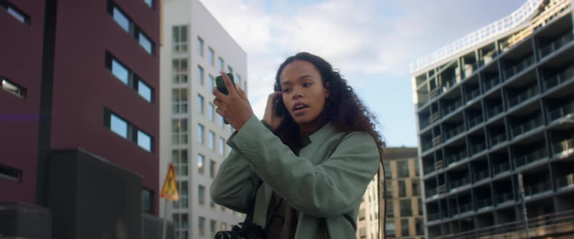 A woman stands outside and holds a cell phone up to fix her hair in the reflection