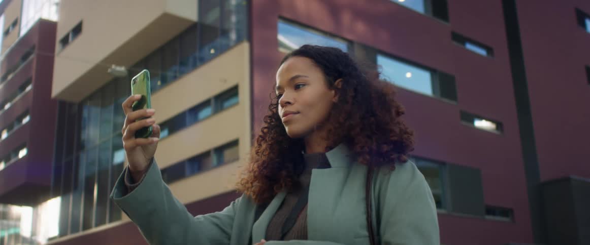 A young woman stands outside an office building and holds up her phone to take a selfie