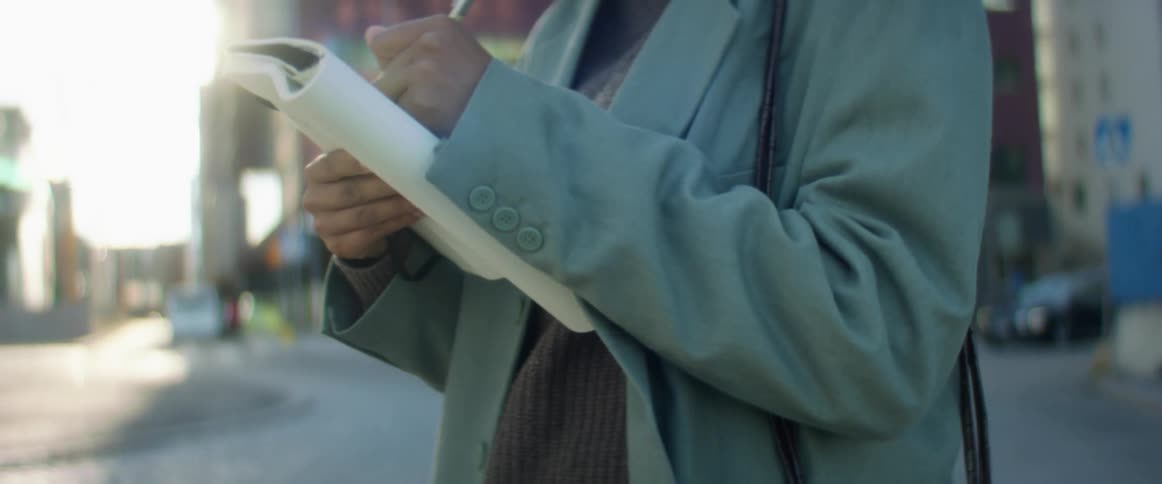 A woman stands outside of large city buildings and writes in a notebook