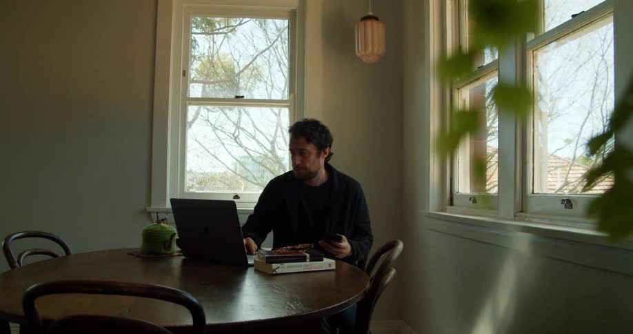 Man with a beard and a black jacket sits at table and boots up a laptop in a room