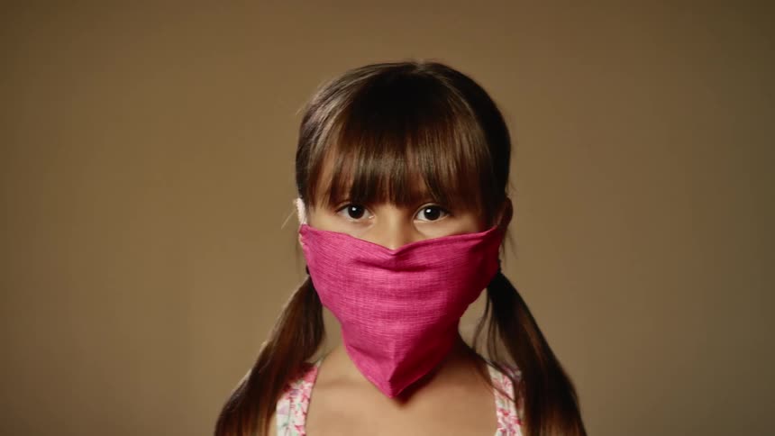 A young girl with brunette pigtails poses while wearing a pink face mask
