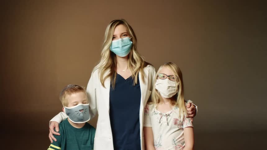 Mother wearing a blue mask puts her hands on her son and daughters shoulders and looks at the camera with eye contact
