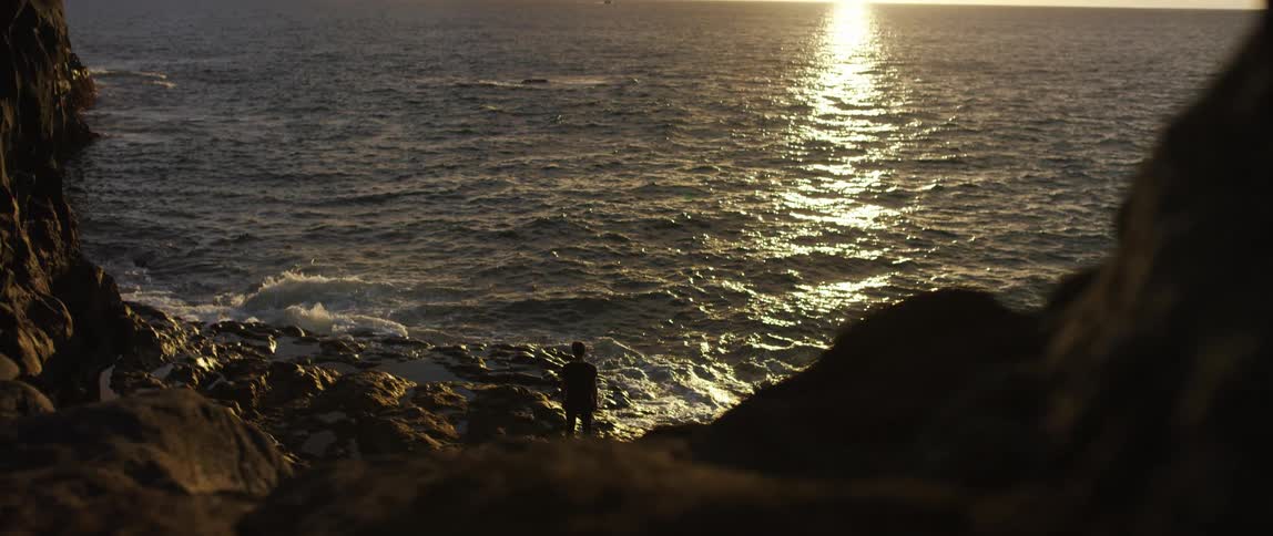 Young man stands on the rocks of a beach on the shoreline next to the ocean at sunset