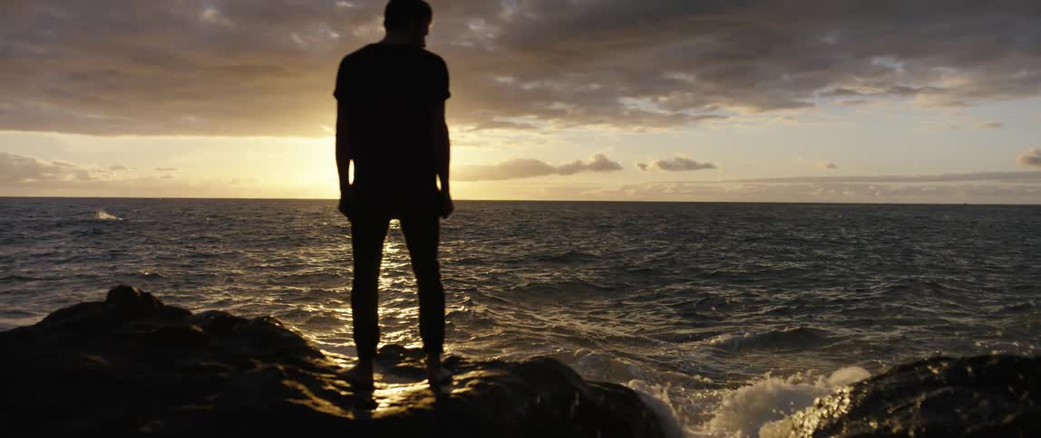 A man standing on rocks on the shore looks out at the water splashing