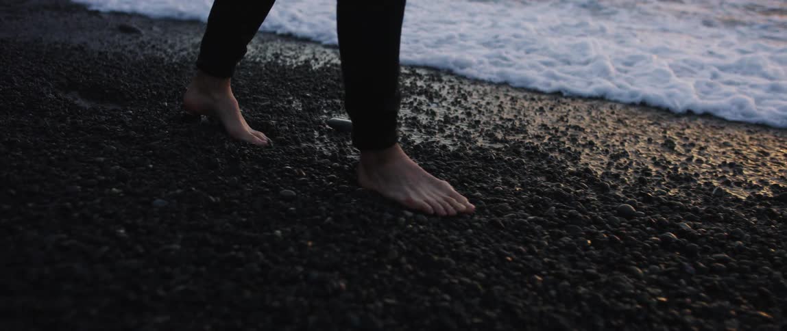 A view of the feet of a man walking barefoot on the pebbles of a beach at dusk