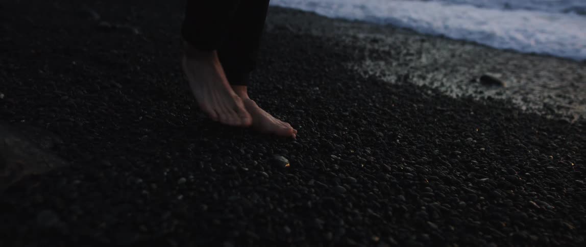 Man with black pants walks barefoot along the beach with pebbles at dusk