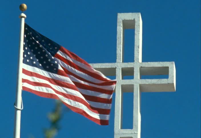 An american flag blows in the wind next to a large hollow cross