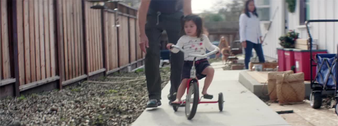 A little girl rides her tricycle in the back yard while her mom watches and her dad helps her