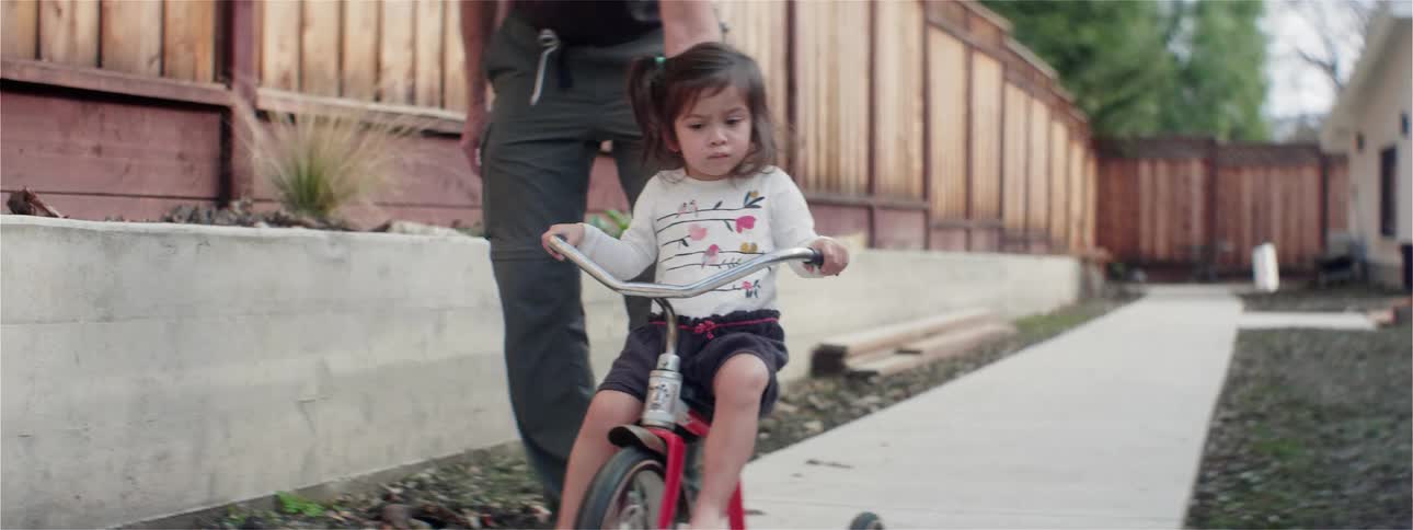 A man pushes his daughter as she rides a tricycle down a sidewalk in their yard