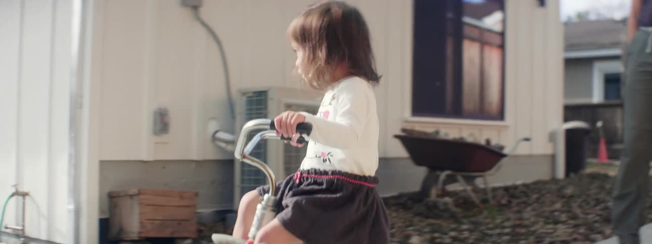 A young girl rides a tricycle around a house as a man watches over her from behind
