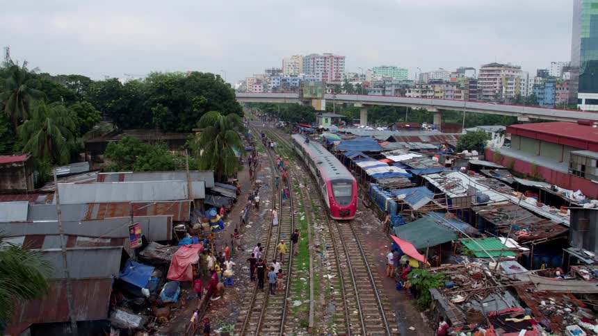 An aerial view of a train driving on tracks through a poor beat up neighborhood as people walk along the tracks