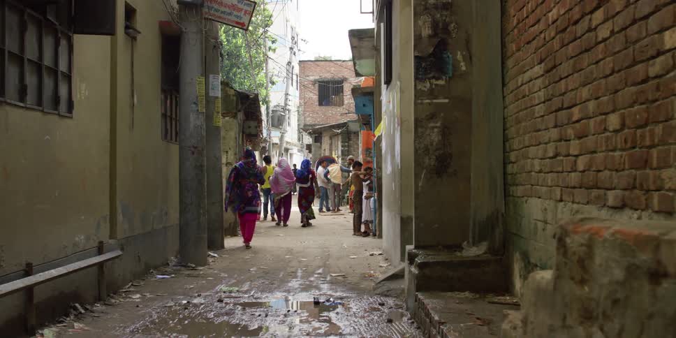 A group of women in colorful clothing and head scarfs walk down a muddy alleyway in a city during the day