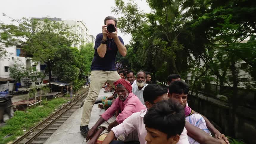 Photographer balances precariously on train car among sitting men as takes pictures