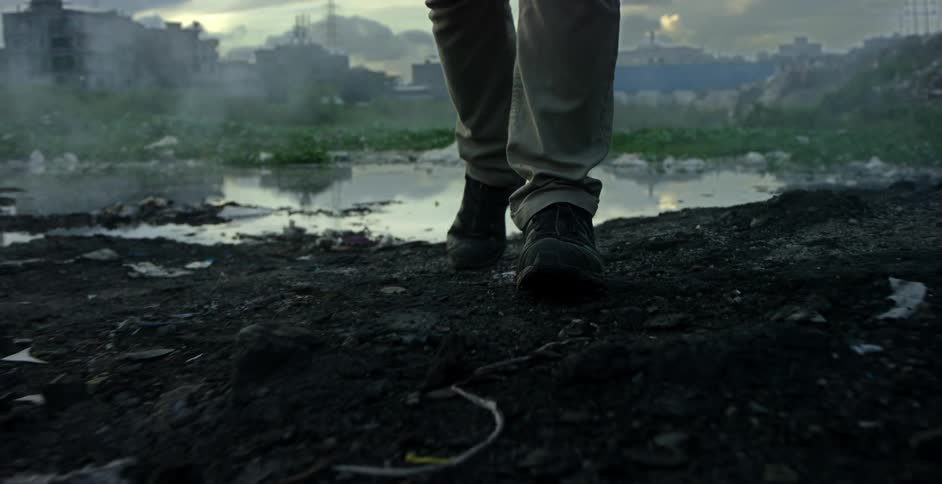 A person wearing work boots and pants walks away from a muddy trashstrewn pond with buildings and power lines in the distance