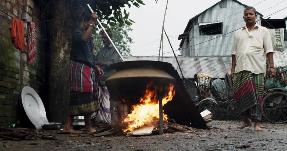 A man uses a pool to stir a large pot over an outdoor fire as another man watches him from nearby