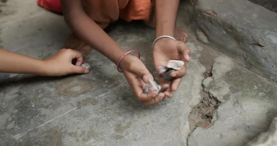 A little girl kneeling outdoors plays with rocks along with a pair of boys