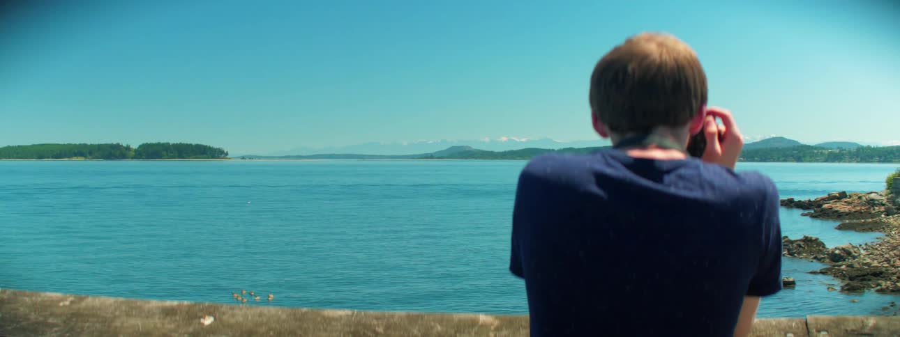 A young adult male stands steadily as he takes aim with his camera at ocean water