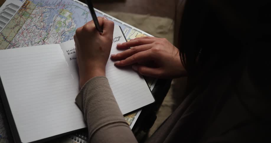 Woman with black hair and a brown sweatshirt writes in a notebook on a map
