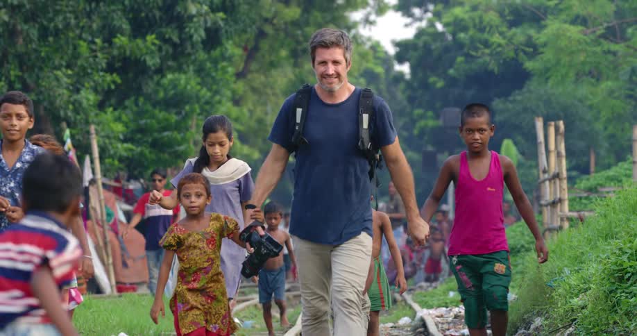 A man carrying a camera holds hands with two children as they walk with a large group of other children in a village