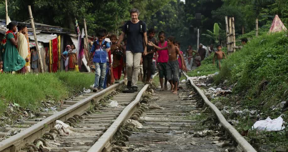 A middle aged caucasian photographer walks atop a train track accompanied by a crowd of ethnic children