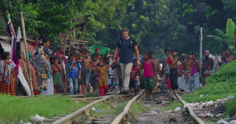 A middle aged caucasion man holding hands with impoverished children walks along a train track as a crowd of children follow behind him