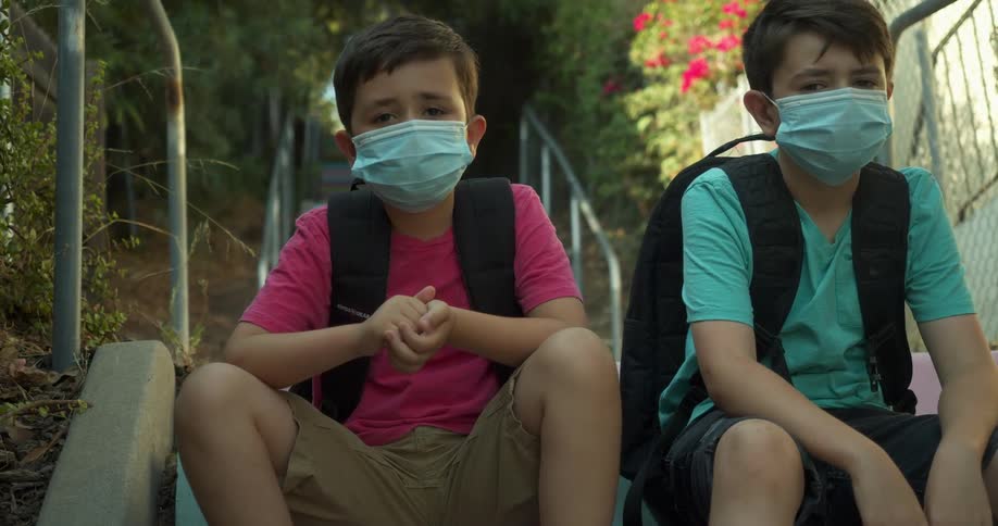 A pair of boys wearing masks and backpacks look into the camera while sitting on a colorful outdoor staircase
